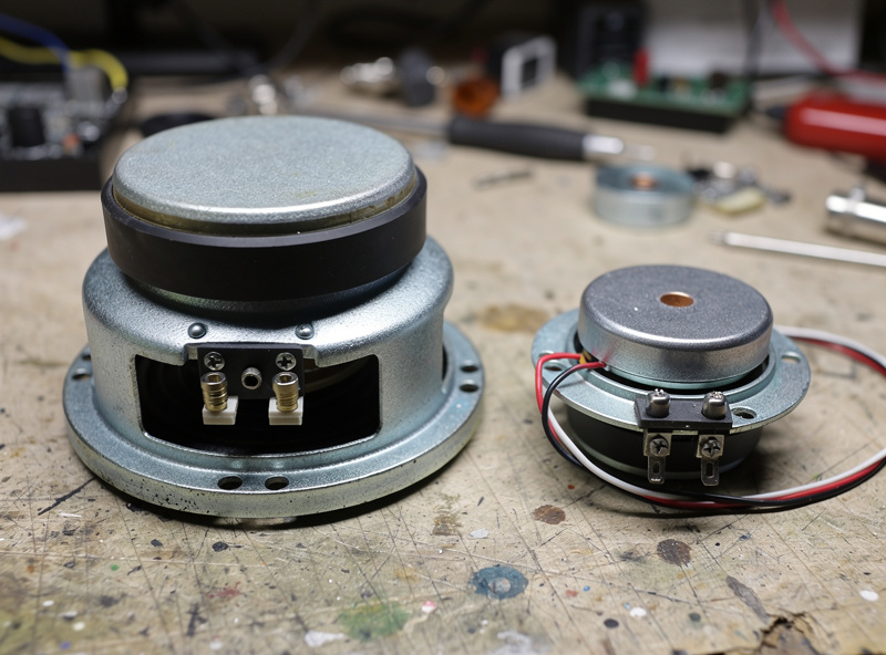 A large bass shaker and a small tactile transducer puck side by side on a workbench