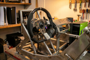 Direct-drive wheelbase mounted to a welded steel cockpit with a PS5 and Xbox console nearby
