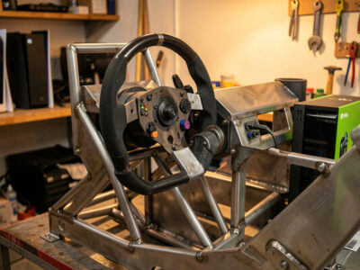 Direct-drive wheelbase mounted to a welded steel cockpit with a PS5 and Xbox console nearby