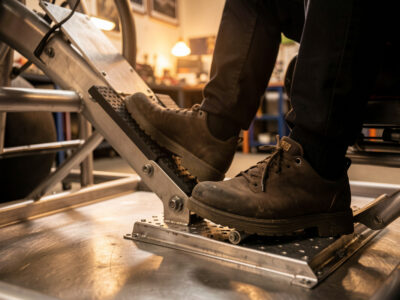 A sim racer's leg pressing a load-cell brake pedal on a welded steel pedal deck