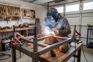 Builder MIG welding a steel-tube sim rig frame in a home garage with clamps holding it square